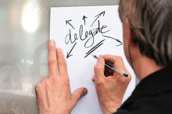 Hand writing the word delegate on a white paper with arrows pointing outward, signifying delegation