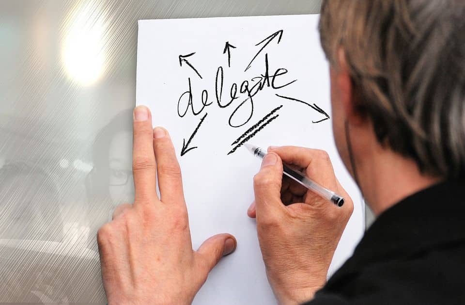 Hand writing the word delegate on a white paper with arrows pointing outward, signifying delegation
