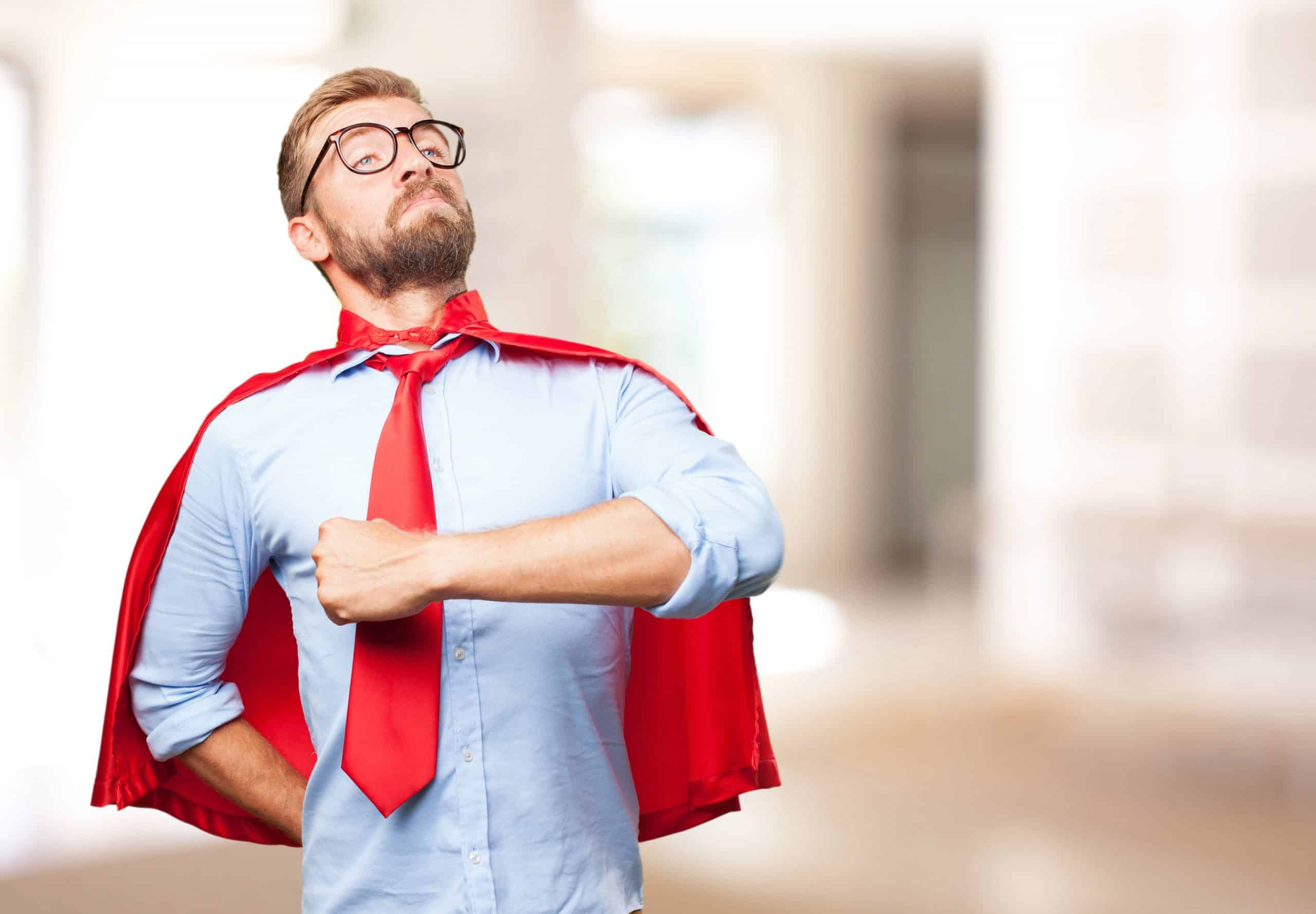 A confident man wearing glasses and a red superhero cape over a blue shirt and red tie, posing with a proud expression in an office setting