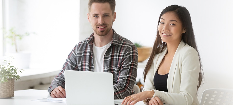 Two people, a man and a woman, sitting at a desk with a laptop. They are both smiling and looking at the camera