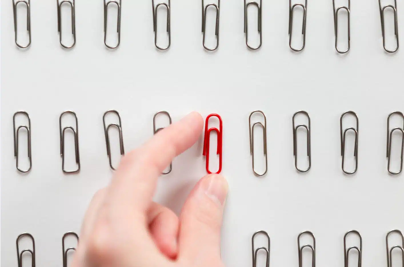 A hand picking up a red paperclip from a row of silver paperclips arranged neatly on a white surface