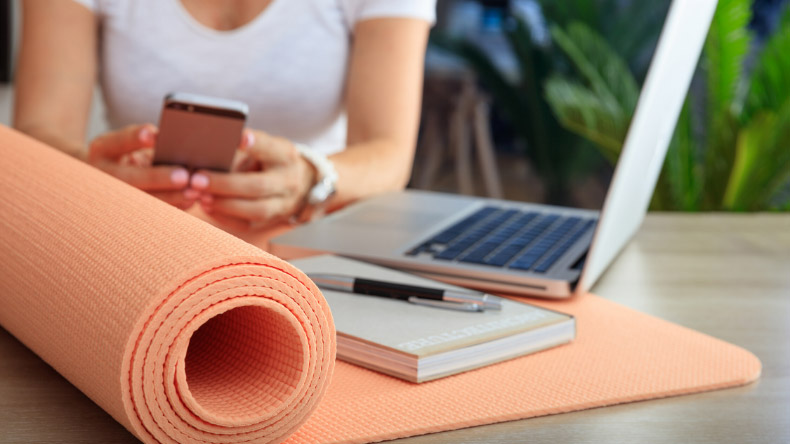 woman holding her cellphone with her laptop, notebook, pen, and yoga mat on the table in front of her
